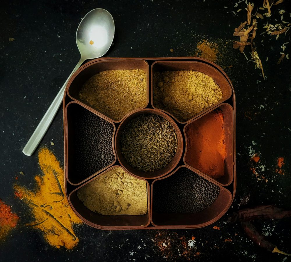 A top view of a spice box filled with various Indian spices, surrounded by scattered colorful powders.